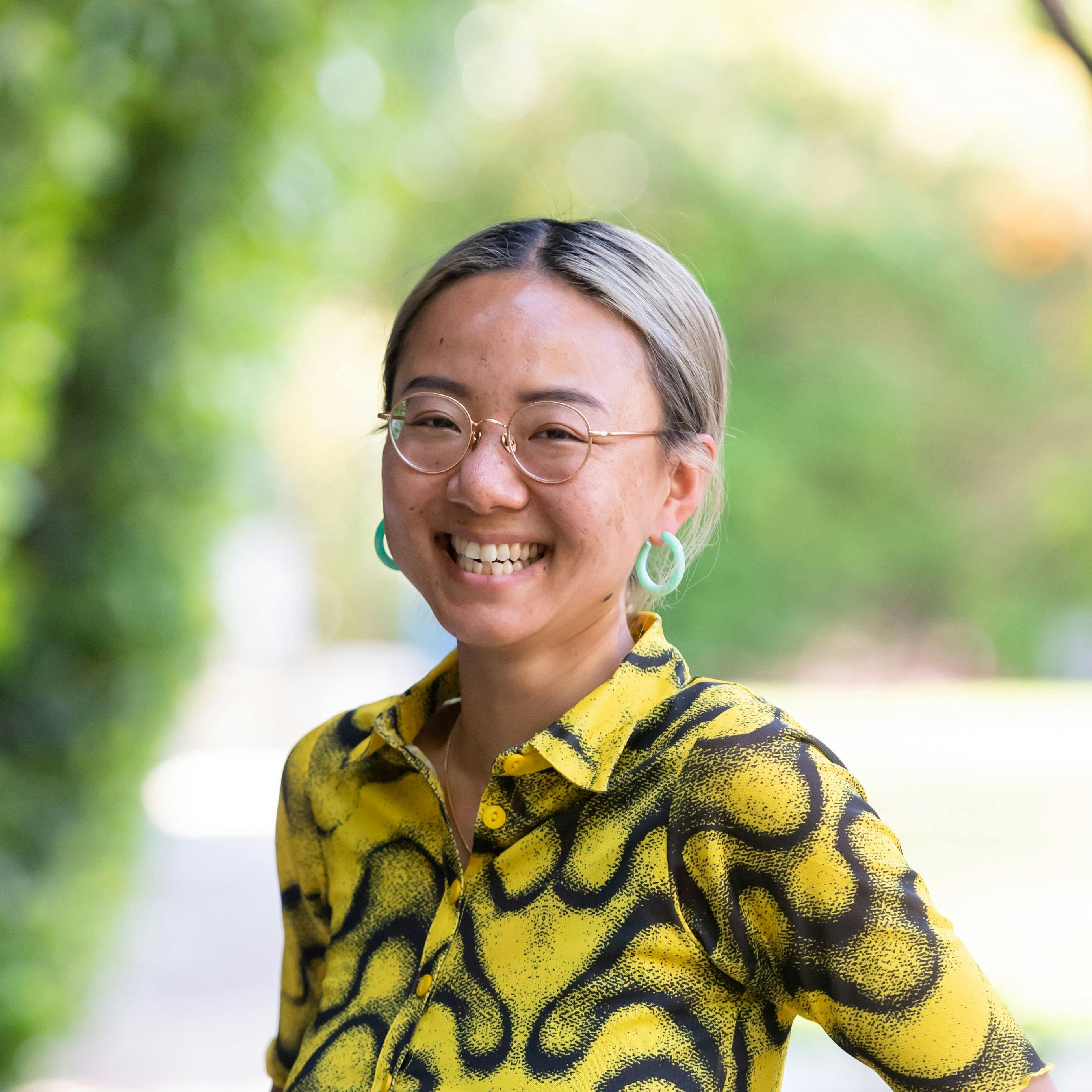 An Asian non-binary person wearing green hoop earrings and a bright yellow shirt with squiggly black lines and dots smiles brightly.