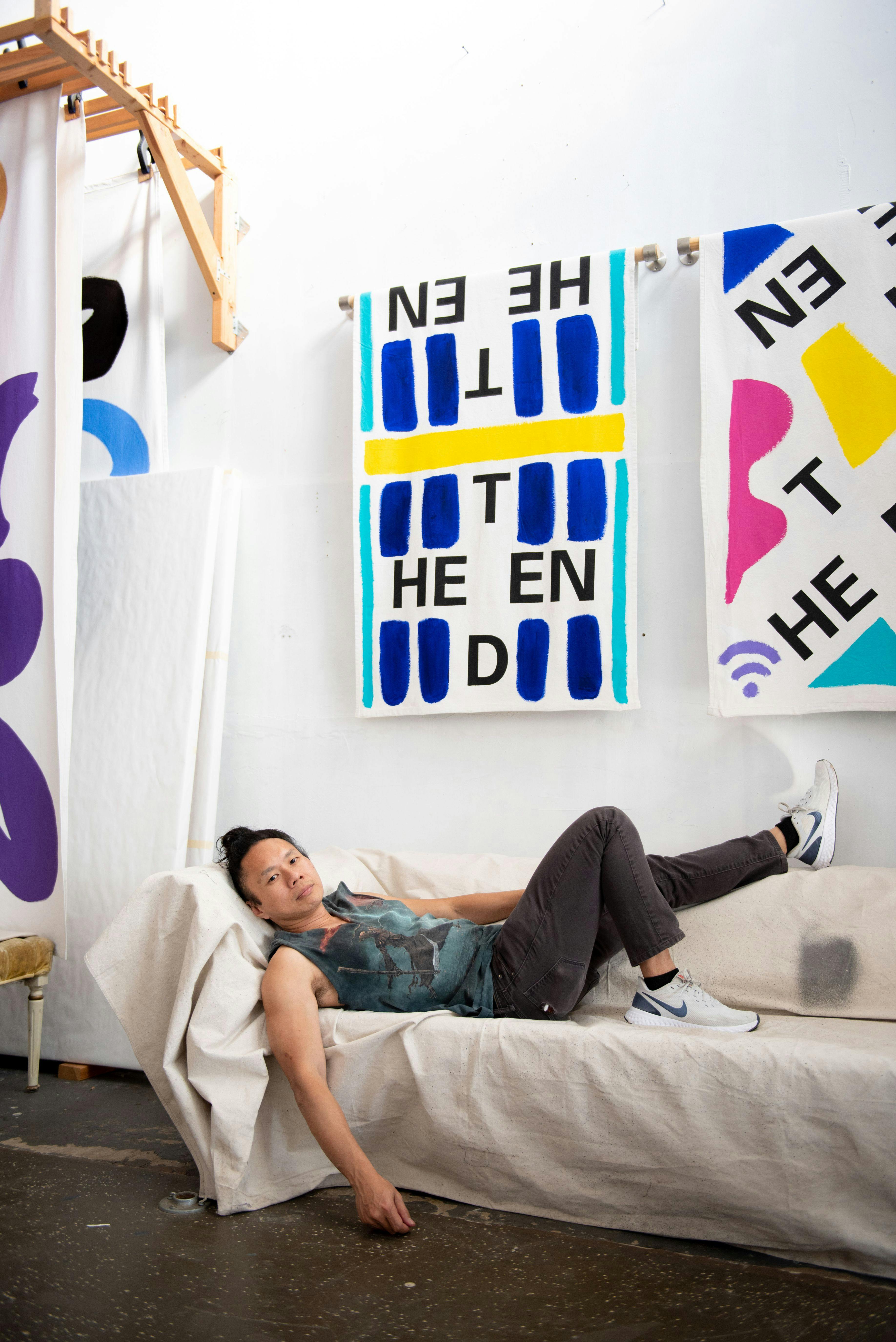 An Asian man wearing black jeans, a graphic teal tank top, and Nikes lies sprawled out on a canvas-covered couch, looking at the camera. Above and behind him are paintings of brightly colored, graphic shapes and letters.