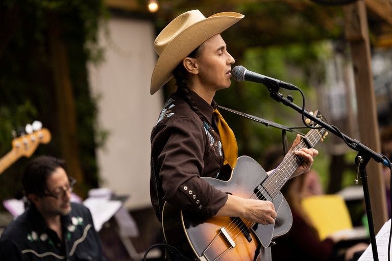 A person seen in profile in an outdoor space with lush vegetation plays guitar and sings into the microphone with their eyes closed. Their hair is in two braids and they wear a cowboy hat, brown vaquero shirt, and a yellow ochre scarf around their neck.