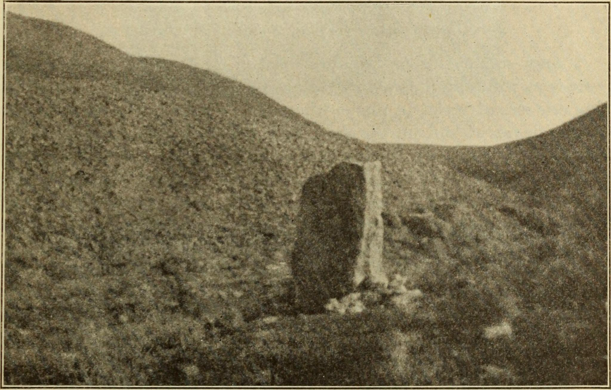 A grainy sepia photograph of rolling hills, foregrounded by a large upright rock.
