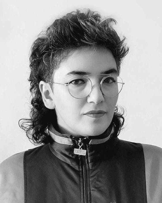 A person with a dark, shoulder-length mullet looks into the camera. She wears wire-frame glasses, a zip-up track jacket, and lipstick.