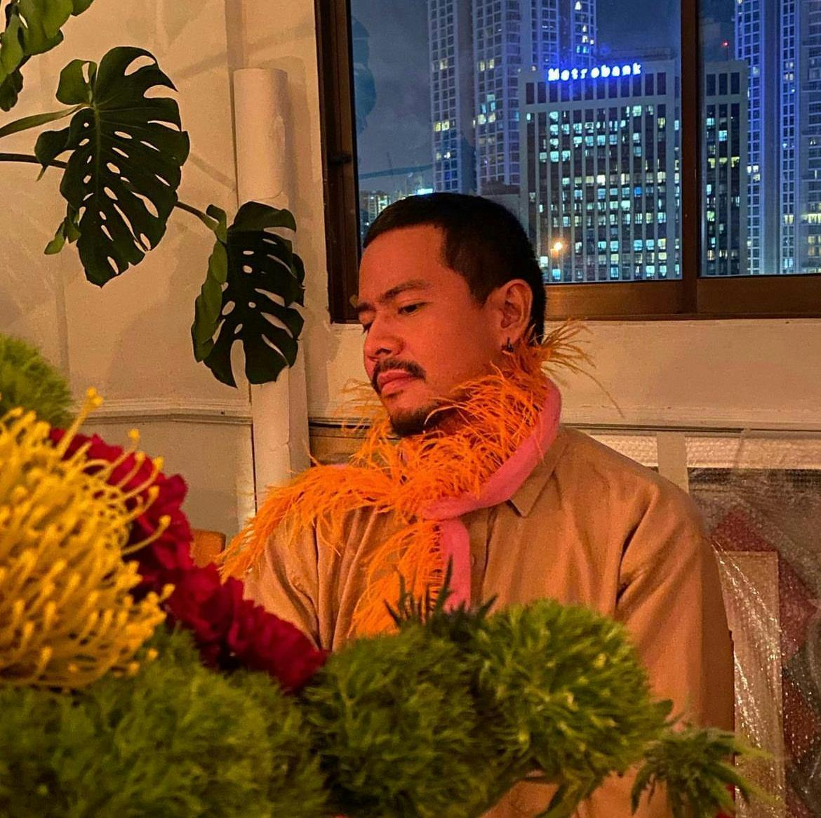 Derek sits in a warmly-lit room, with a view of a city at night outside of the window. He looks down at a tabletop of large flowers and lush greenery and wears a feathery boa around his neck.